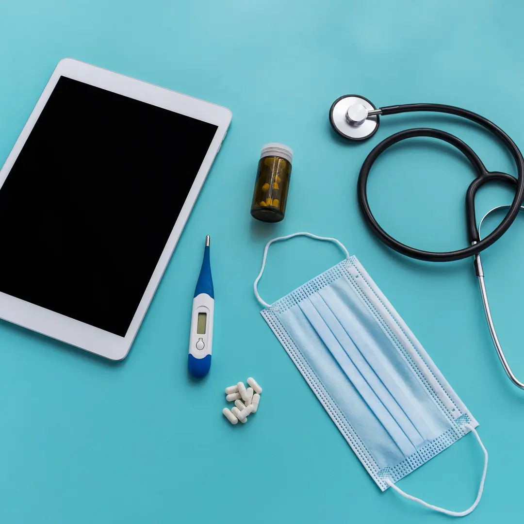 Tablet, disposable mask, stethescope, rx bottle, pills on a blue table.