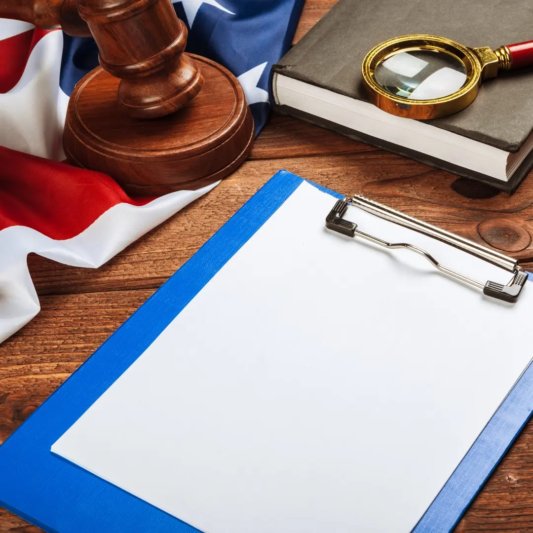 Image of a clipboard, magnifying glass on a book, gavel and an American flag on a wood desk.