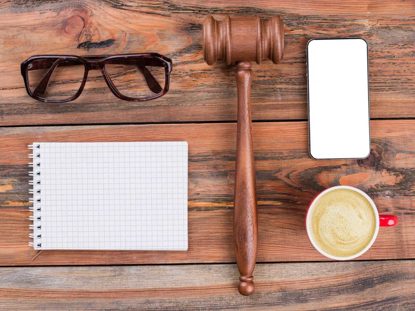 High resolution photo of a notepad, eyeglasses, gavel, phone and a cup of coffee in a red cup on a wooden table.